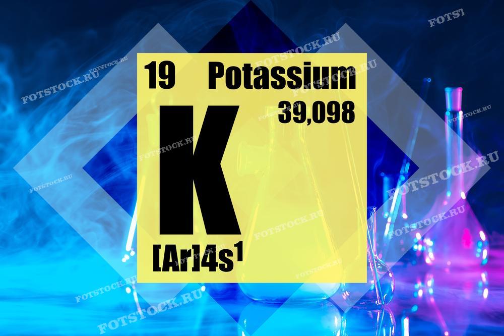 На фотографии изображены надпись "Potassium", химическая формула калия ([Ar]4s¹) и его атомный номер 19 на желтом фоне. На заднем плане видны лабораторные колбы и пробирки, освещенные разноцветными огнями, создающие атмосферу научных исследований в лаборатории.